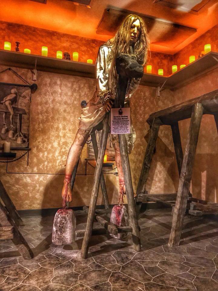 a medieval torture exhibit featuring a mannequin depicting a person tied to a wooden contraption, suspended in a distressing posture. The dimly lit room, with stone-textured flooring and warm lighting, adds to the eerie atmosphere.