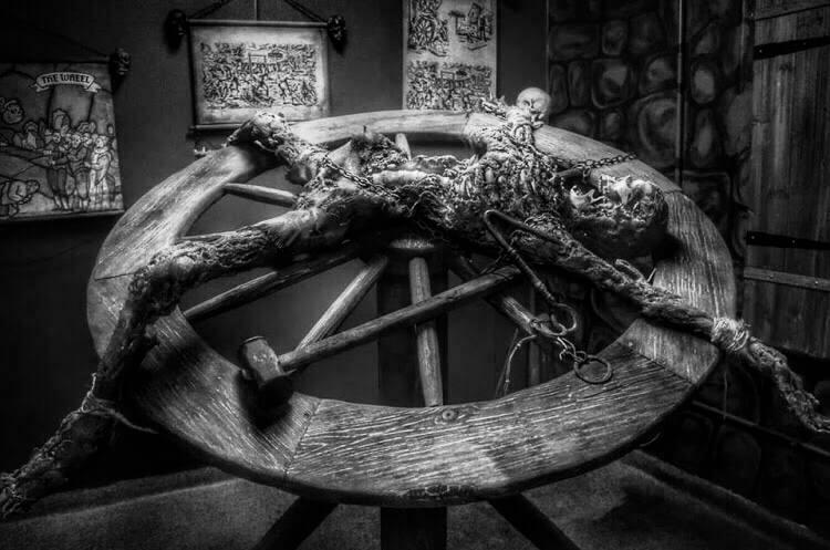 a gruesome exhibit of a medieval torture device known as the breaking wheel. A life-sized mannequin, depicting a decayed and tortured figure, is bound and stretched across the large wooden wheel, with chains and restraints holding its limbs in place.