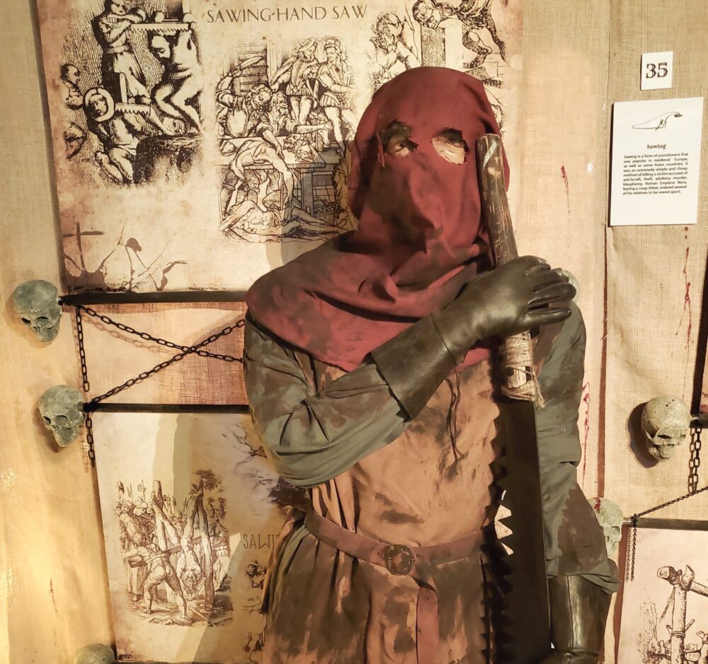 a person or mannequin dressed in medieval-style clothing, wearing a red hood and leather gloves, possibly portraying a torturer or executioner, with old illustrations in the background, likely part of an exhibit related to historical torture methods.