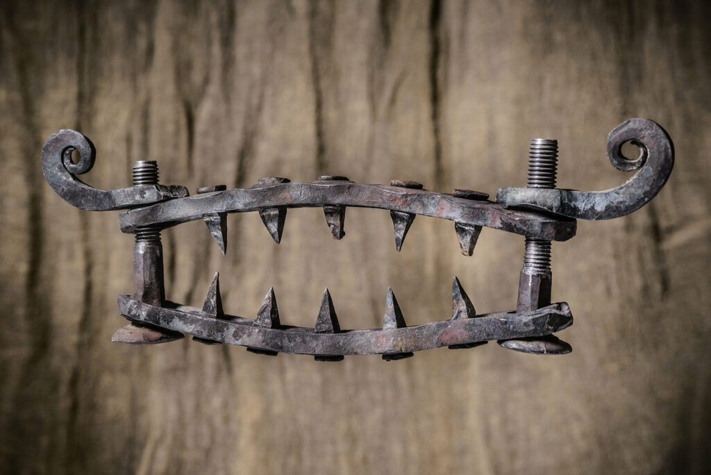 an iron device with sharp, pointed spikes and screws, resembling a jaw-like trap. This appears to be an instrument of torture or punishment, possibly designed to cause pain by tightening the screws and pressing the spikes into the victim's body.