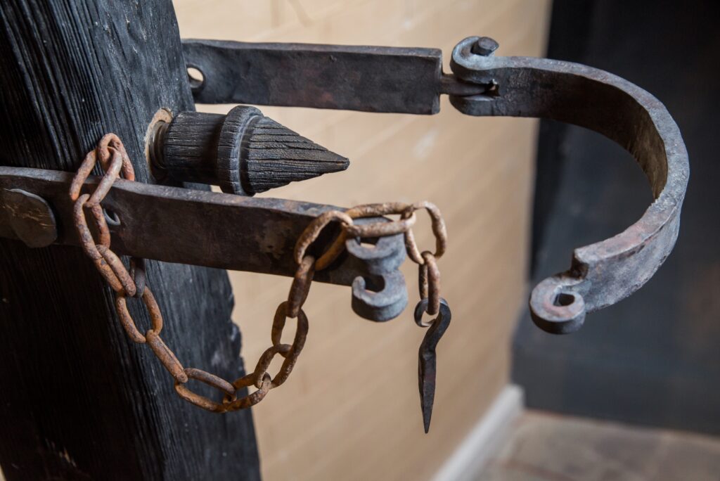 a rusted medieval-style metal shackle, featuring a large spike and an attached chain. It appears to be a part of a restraint device, likely used for locking prisoners or as part of torture instruments.