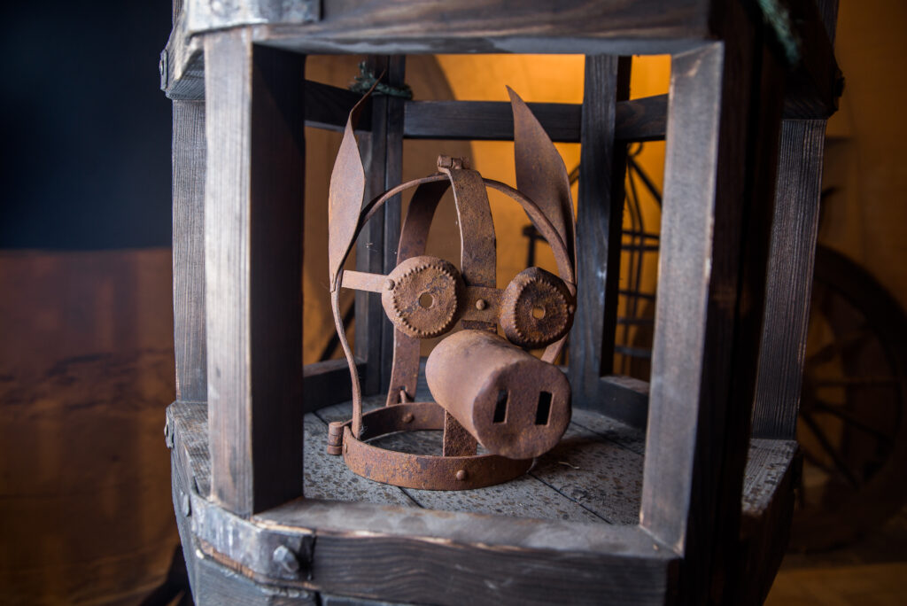 a metal torture device resembling a mask with animal-like features, possibly a pig. It is displayed within a wooden cage or frame.