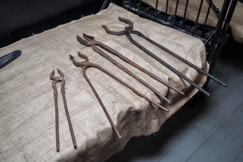 a collection of metal tools, likely tongs or pincers, laid out on a burlap-covered surface. These tools appear aged and could have been used historically for torture or manual labor.