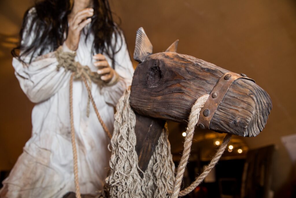 a mannequin dressed in historical clothing with tied hands, standing beside a wooden horse figure.