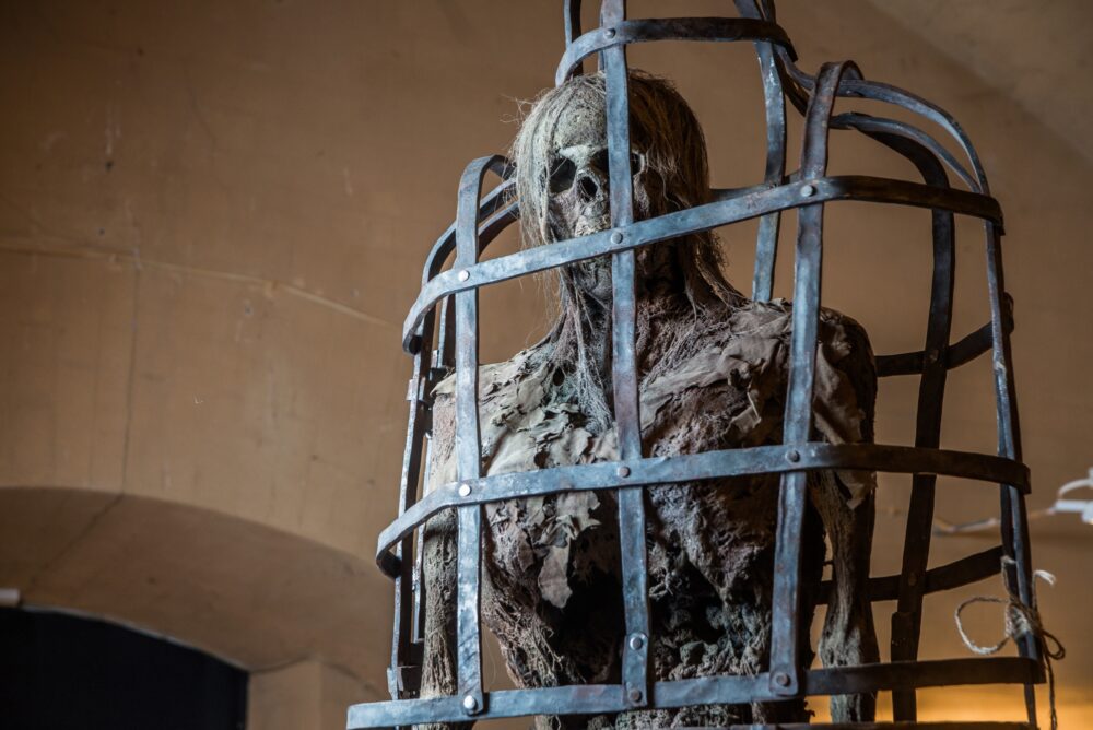 a skeleton or mummified figure enclosed in an iron cage, evoking a dark, historical or medieval theme, likely related to torture or punishment displays in a museum setting.