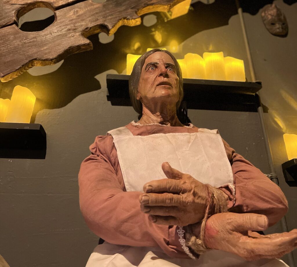 a lifelike mannequin depicting a historical scene. The figure appears to represent a bound woman in a distressed state, possibly linked to a historical event such as witch trials or other forms of persecution.