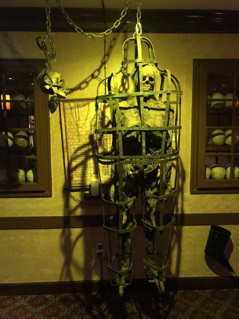 skeletal figure enclosed in a metal cage, suspended by chains, displayed in a dimly lit room with an eerie atmosphere