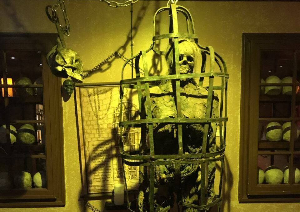 skeletal figure enclosed in a metal cage, suspended by chains, displayed in a dimly lit room with an eerie atmosphere