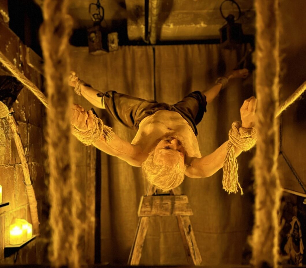 lifelike figure of a man suspended by ropes, arms and legs tied, in a dimly lit room with a medieval, dungeon-like ambiance