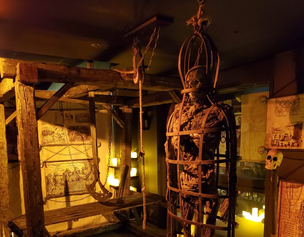 a dimly lit medieval torture museum exhibit featuring a life-sized human figure enclosed in an iron cage, resembling the historical practice of gibbetting