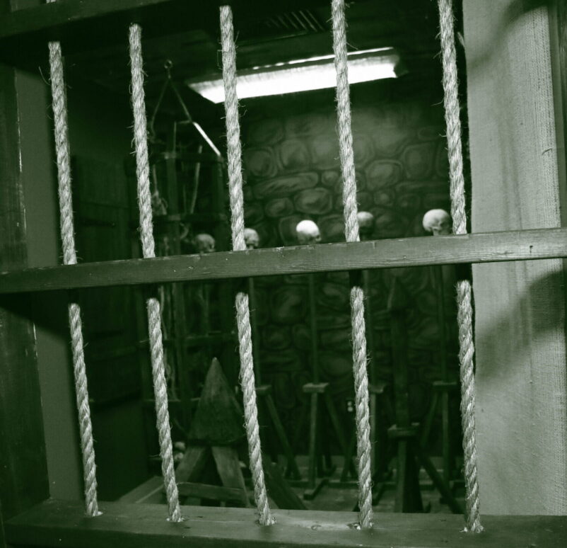 a view through a barred window, revealing a dark, eerie room filled with torture devices and skulls.